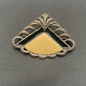 May include: A silver tone brooch with a black and cream enamel design. The brooch is shaped like a triangle with a scalloped edge.