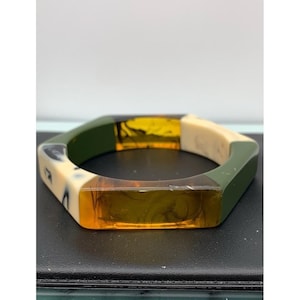 May include: A geometric bangle bracelet with olive green, cream, and amber-colored segments. The bracelet has a hexagonal shape and is made of a translucent material. The amber sections have a marbled effect.