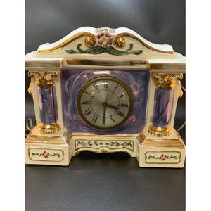 May include: A decorative ceramic clock with a cream-colored base and gold accents. The clock face is silver with black Roman numerals. The clock features purple columns and floral details, with a decorative top.