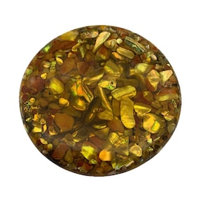May include: A round, decorative object filled with a mix of golden, brown, and iridescent pieces. The pieces are embedded in a clear, resin-like material, creating a textured and visually appealing surface. The object is set against a white background.