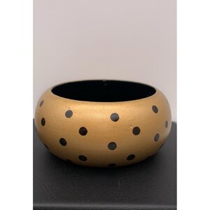 May include: A wide, circular bangle bracelet featuring a metallic gold exterior surface decorated with a repeating pattern of small, solid black polka dots. The interior of the bangle is finished in a solid black color, creating a contrast.