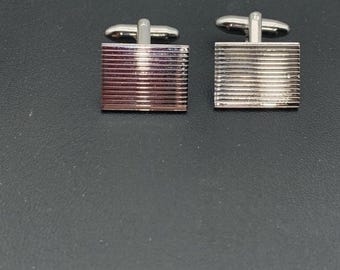 May include: A pair of silver-toned cufflinks with a rectangular face featuring horizontal lines. The cufflinks have a T-bar closure and are displayed on a dark surface. The design is simple and elegant.