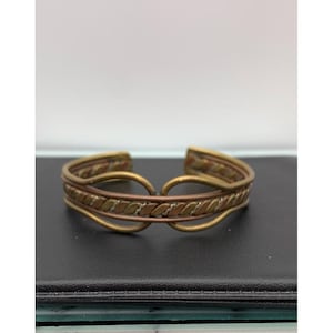 Op de afbeelding: Een vintage manchetarmband met een gemengd metalen ontwerp. De armband heeft een centrale band van koperkleurig metaal met een herhalend bladerpatroon, omlijst door goudkleurig metaal. Het open ontwerp maakt het gemakkelijk om te dragen.