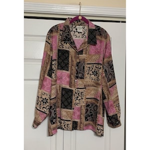 May include: A long-sleeved button-down shirt featuring a patchwork pattern in shades of pink, black, and tan. The design includes floral motifs, paisley swirls, and geometric diamond patterns. The shirt has a classic collar and a front button closure.