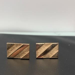 May include: A pair of gold-toned rectangular cufflinks. Each cufflink features a diagonal striped design. The cufflinks are displayed on a dark surface, with a plain white background.