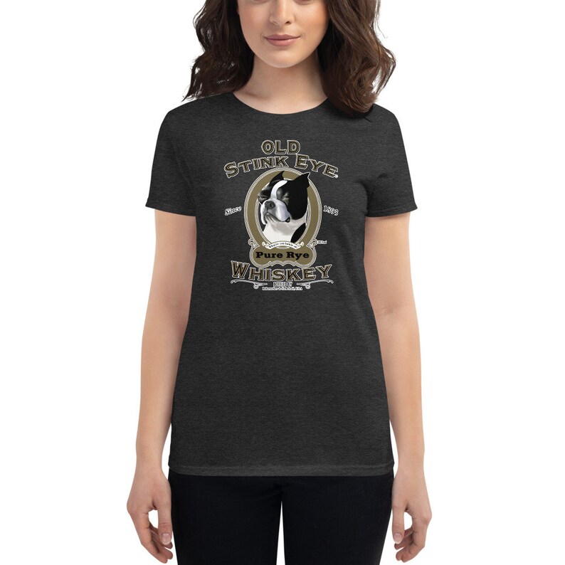 May include: A dark heather grey women's t-shirt with a vintage-style illustration of a Boston Terrier dog. The illustration is surrounded by a gold oval frame with the text "Old Stink Eye Pure Rye Whiskey" in a stylized font.