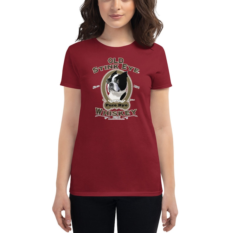 May include: A red women's t-shirt with a vintage-style illustration of a Boston Terrier dog. The illustration is surrounded by a gold oval and the text "Old Stink Eye Pure Rye Whiskey".