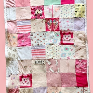 May include: A pink, white, and cream patchwork quilt with a variety of fabric patterns and textures. The quilt features squares with Minnie Mouse, diamond shapes, and the word "HAPPY". It has a white border around the edges.