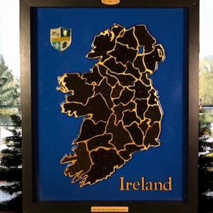 Könnte beinhalten: Gerahmtes Kunstwerk mit einer Karte von Irland mit goldfarbenen Umrissen auf blauem Hintergrund. Das Wort "Ireland" ist in goldfarbenen Buchstaben geschrieben. Ein kleines Emblem befindet sich in der oberen linken Ecke. Der Rahmen ist schwarz.