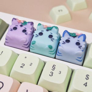 Op de afbeelding: Drie kleurrijke katvormige toetsdoppen in lavendel, mintgroen en blauw, elk met een bloemaccent. De toetsdoppen staan op een wit toetsenbord, met lichtgroene en witte toetsen zichtbaar. Deze katten-toetsdoppen geven elk toetsenbord een speelse uitstraling.