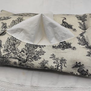 Puede incluir: Una funda rectangular para caja de pañuelos con un estampado toile de Jouy en blanco y negro que representa árboles, figuras y pájaros. Los pañuelos blancos son visibles a través de la abertura. La funda es de tela.