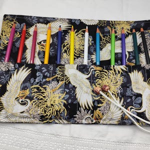 Peut inclure: Un rouleau à crayons en tissu noir avec un motif de grue blanche et des accents dorés. Le rouleau comporte 12 fentes pour crayons et une fermeture à cordon blanc.