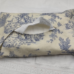 May include: A rectangular cushion featuring a blue and white toile de Jouy design. The fabric shows scenes of figures, trees, and birds. The cushion has a white interior and a folded flap.