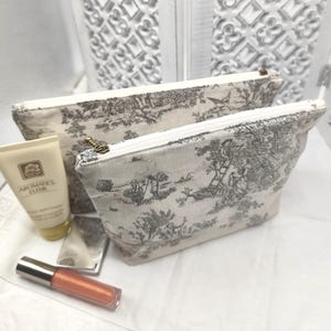 Peut inclure: Deux pochettes zippées beiges avec un motif toile noir et blanc. Les pochettes sont en tissu et ont une fermeture éclair. La plus petite pochette est à l'intérieur de la plus grande.