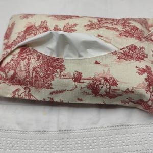 Peut inclure: Un étui à mouchoirs rectangulaire avec un motif de toile de Jouy rouge sur fond crème. Le tissu représente des scènes pastorales avec des personnages, des arbres et des bâtiments. L'ouverture révèle un intérieur blanc.