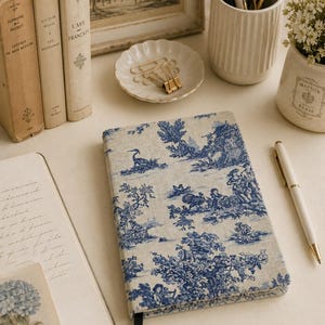 Può includere: Un taccuino rettangolare con copertina in tessuto beige e motivo toile de jouy blu che raffigura scene pastorali con figure, uccelli e alberi. Il taccuino è dotato di un segnalibro a nastro nero ed è posizionato accanto a una penna bianca.