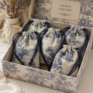 Peut inclure: Une boîte rectangulaire contenant cinq petits sachets en tissu à cordon, tous ornés d'un motif toile de jouy bleu et crème. L'intérieur du couvercle porte l'inscription SACHETS PARFUMÉS. Les sachets sont fermés par des cordons bleu foncé.