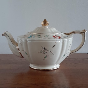 May include: Cream-coloured ceramic teapot with a gold handle, spout, and trim. The teapot features a floral design with red, blue, and green accents. The lid is topped with a gold finial. The teapot is on a wooden surface.
