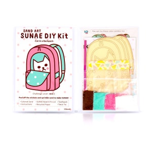 May include: A pink and blue backpack with a cat peeking out of the pocket. The kit includes coloured sand, a pre-cut cardboard backpack, a toothpick, and a twist tie. The text on the packaging reads "SAND ART SUNAE DIY Kit -Cat in a Backpack- Challenge Level: ★★☆ Peel off the stickers and sprinkle sand to make SUNAE! •Coloured Sand SUNAE Board (Pre-cut) Toothpick -Instructions *Recycled Paper •Twist Tie ©Naoshi"