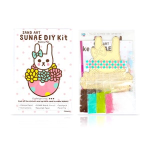May include: A DIY sand art kit featuring a bunny in a pot with flowers. The kit includes coloured sand, a pre-cut board, a toothpick, and a twist tie. The kit is labelled "SUNAE DIY Kit - Bunny Pot -" and has a challenge level of three stars.