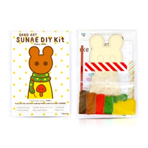 May include: A sand art DIY kit featuring a cartoon bear with a red mushroom and a green snake. The kit includes a wooden bear cutout, coloured sand, a toothpick, and a twist tie. The packaging is clear plastic and shows the contents of the kit.