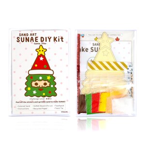 May include: A Christmas-themed sand art kit featuring a cartoon Santa Claus tree design. The kit contains coloured sand, a pre-cut board, a toothpick, a twist tie, and instructions. The kit is packaged in a clear plastic box.