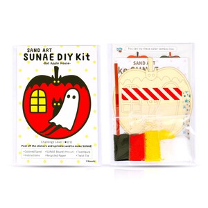 May include: A red apple-shaped wooden craft kit with a ghost and a black cat inside. The kit includes coloured sand, a pre-cut wooden board, a toothpick, and a twist tie. The text on the packaging reads "Sand Art SUNAE DIY Kit -Bat Apple House- Challenge Level:  Peel off the stickers and sprinkle sand to make SUNAE!"