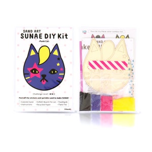 May include: A sand art kit featuring a punk cat design. The kit includes a pre-cut wooden board, coloured sand, a toothpick, and a twist tie. The packaging is pink and white with the text "SAND ART SUNAE DIY Kit -Punk Cat-  Challenge Level: ★★☆ Peel off the stickers and sprinkle sand to make SUNAE! •Colored Sand •SUNAE Board (Pre-cut) •Toothpick •Instructions •Recycled Paper •Twist Tie ©Chaeshi"
