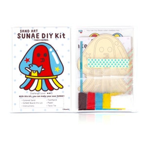 May include: A sand art DIY kit featuring a cartoon jellyfish character named "SUNAE". The kit includes coloured sand, a pre-cut wooden board, a toothpick, paper, and a twist tie. The packaging is white with blue and red accents.