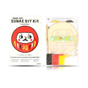 May include: A Sand Art Sunae DIY Kit with a Lucky Daruma design. The kit contains coloured sand, a pre-cut Sunae board, instructions, a toothpick, recycled paper, and a twist tie. The packaging is white with red, yellow, and black accents.