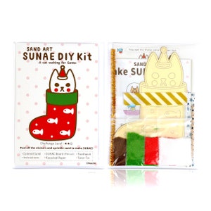 May include: A white cat wearing a Santa hat is peeking out of a red stocking with white fish designs. The cat is part of a sand art DIY kit that includes coloured sand, a pre-cut board, a toothpick, and a twist tie. The kit is labelled "SAND ART SUNAE DIY Kit - A cat waiting for Santa -"