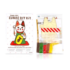 May include: A sand art DIY kit featuring a calico cat with a fish. The kit includes coloured sand, a pre-cut board, a toothpick, and a twist tie. The kit is labelled "SUNAE DIY Kit -Lucky Calico-."