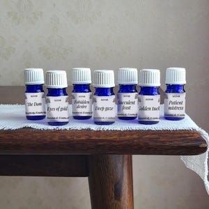 Könnte beinhalten: Sechs kleine blaue ätherische Ölflaschen mit weißen Kappen sind auf einem weißen Tuch angeordnet. Jede Flasche hat ein Etikett mit einem anderen Namen: "The Dom", "Eyes of gold", "Forbidden desire", "Deep gaze", "Succulent feast" und "Patient mistress".