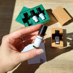 May include: A close-up of a small brown glass bottle with a black rollerball top. The bottle is labeled "Sunrise" and is part of a set of essential oil roll-ons.