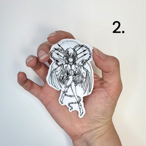 Op de afbeelding: Zwart-wit sticker met een gedetailleerde illustratie van een gevleugelde figuur met kettingen. De sticker wordt in een hand gehouden tegen een witte achtergrond, met het nummer "2" zichtbaar.