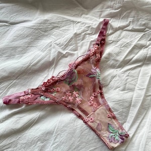 Puede incluir: Un tanga rosa transparente con bordados florales en tonos rosa, verde y morado. La delicada lencería presenta un borde festoneado y tirantes ajustables, sobre una sábana blanca.