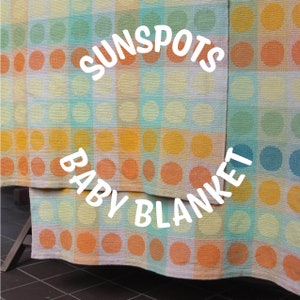 Op de afbeelding: Een babydeken met een geruit patroon van geel, oranje, blauw en groen. De deken heeft een wit cirkelpatroon en de woorden "Sunspots Baby Blanket" staan in wit op de deken.