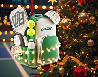 May include: A Christmas ornament shaped like a pickleball bag, racket, and balls. The bag is white and green with the name "Susan" on it. A white hat with a green "S" sits on top. The ornament is decorated with a string of lights and a red ribbon.