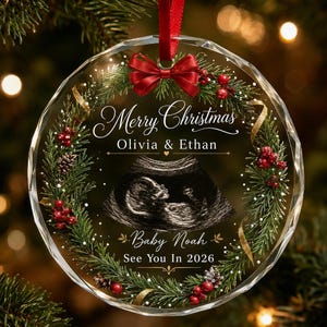 May include: A circular clear glass Christmas ornament featuring a printed wreath of green pine needles, red berries, and pinecones. The center displays an ultrasound image with the text Merry Christmas, Olivia & Ethan, Baby Noah, See You In 2026.