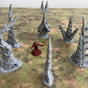 May include: A tabletop gaming scene featuring several gray, spiky terrain pieces and a miniature figure. The terrain pieces vary in shape, resembling twisted trees or alien structures. The miniature figure is red and stands on a red base, adding a pop of color.