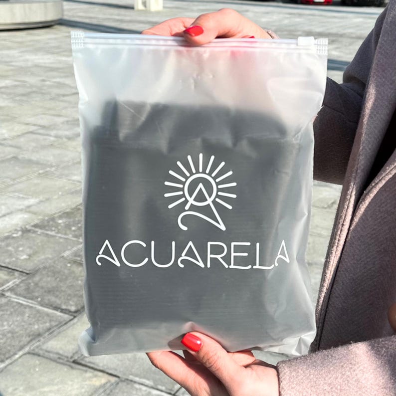 May include: A clear plastic bag with a zipper closure, containing a black item with a white logo that says "ACUARELA" and a stylized sun and mountain design.