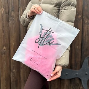 May include: A clear plastic bag with a zipper closure. The bag has a black graphic of a skeleton hand with the text "silk vine" and "silkvineclub.com". The bag is filled with a pink knitted item.