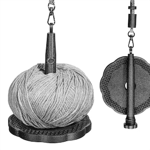 Può includere: Un porta-filo con una palla di filo grigio. La palla di filo poggia su una base nera, strutturata. Un'asta di metallo con una parte superiore decorativa è inserita nella palla di filo. È visibile anche un secondo porta-filo.