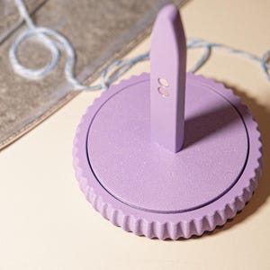 Può includere: Un timbro sigillo color lavanda con base rotonda e bordo scanalato. Il timbro ha un manico rettangolare con due piccoli fori. Sullo sfondo un cordino blu e bianco.