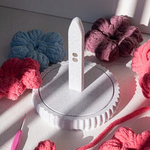 Può includere: Porta fiori in crochet bianco con una base testurizzata e un alto palo bianco con due punti dorati. Il supporto è circondato da fiori all'uncinetto colorati in rosa e blu.