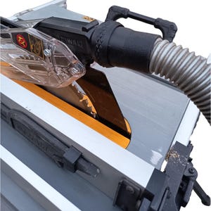 Op de afbeelding: Een close-up van een DeWalt tafelzaag. De zaag heeft een zilveren tafel, een zwarte motorbehuizing en een transparante zaagbladbescherming. Het zaagblad is zichtbaar en er is een stofafzuigslang bevestigd. De zaag heeft een geel en zwart accent.