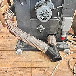 Pode incluir: Uma grande máquina industrial cinza escuro com uma mangueira flexível e canelada. A máquina tem uma ventoinha, componentes elétricos e é montada sobre rodas. A mangueira curva-se para um bocal preto em forma de Y. A máquina está num chão de madeira.