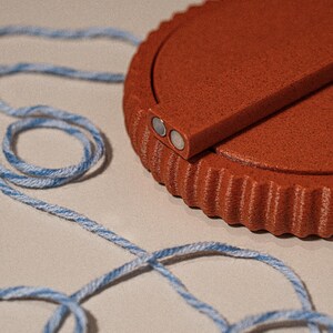 Può includere: Un oggetto rotondo color terracotta con un bordo scanalato e un pezzo rettangolare attaccato, con due piccoli magneti argentati. Un cordoncino ritorto blu e bianco è avvolto attorno all'oggetto.