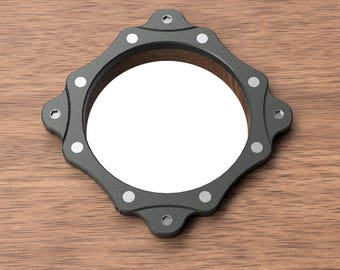May include: A dark gray, geometric-shaped mirror with a circular interior. The frame has a textured appearance and is adorned with small, silver-colored accents. The mirror is set against a wooden surface, creating a contrast in textures and tones.
