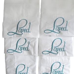 May include: Six white pillowcases with the word "Loved" embroidered in teal on each pillowcase.